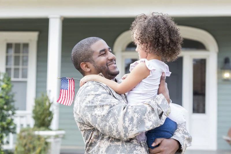 An army dad carrying her child and beside them is their newly bought home