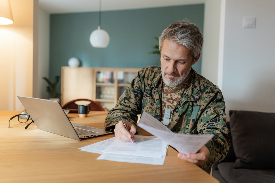 A middle-aged white male military veteran with gray hair and beard sits indoors at a wooden table reviewing documents. Casual home environment with a laptop and shelves in the background