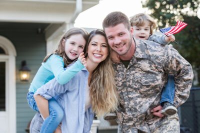 Soldier in uniform gives children piggyback rides outside their home with American flags.